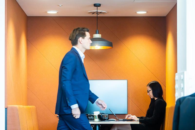 A man in a blue suit walks past an orange seated area in an office, while a woman works on a laptop at the desk